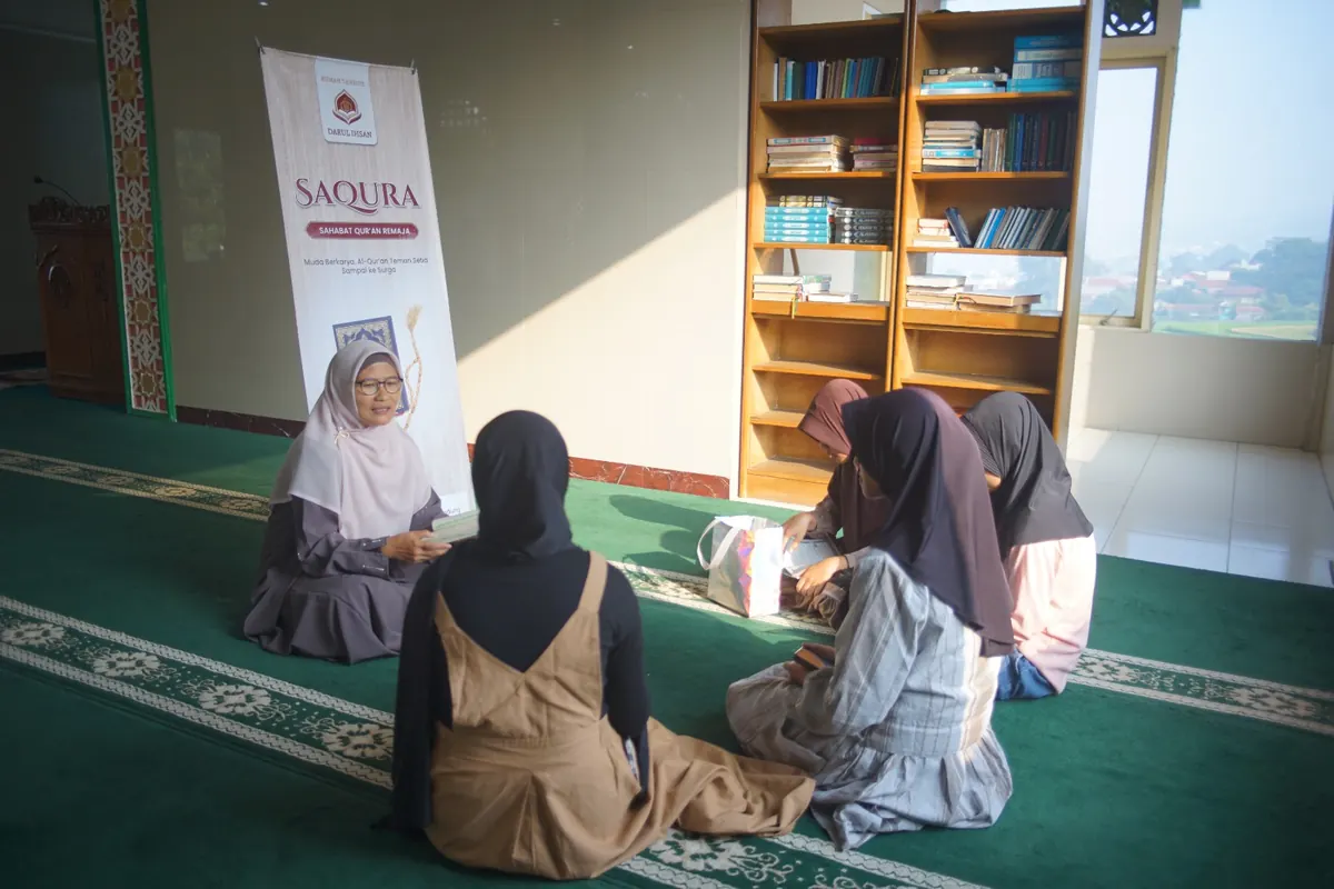 Seorang pengajar berhijab duduk di lantai berhadapan dengan beberapa santriwati di depan rak buku